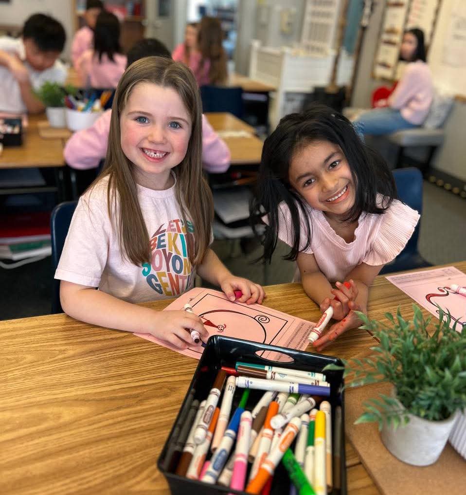 Two young girls smiling at a desk during a coloring activity
